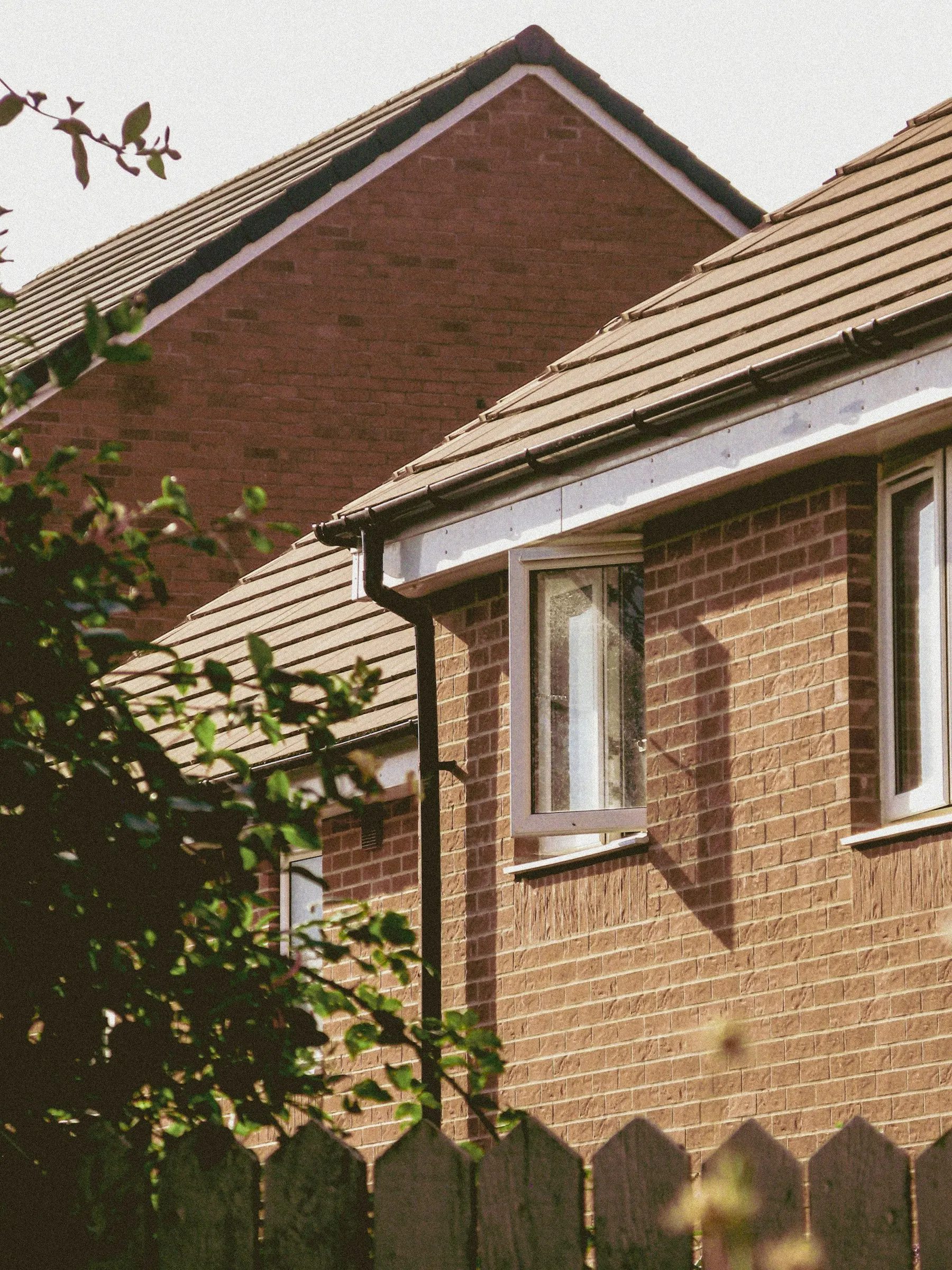 A brick house with a tiled roof, white fascia and a black downpipe, in sunlight