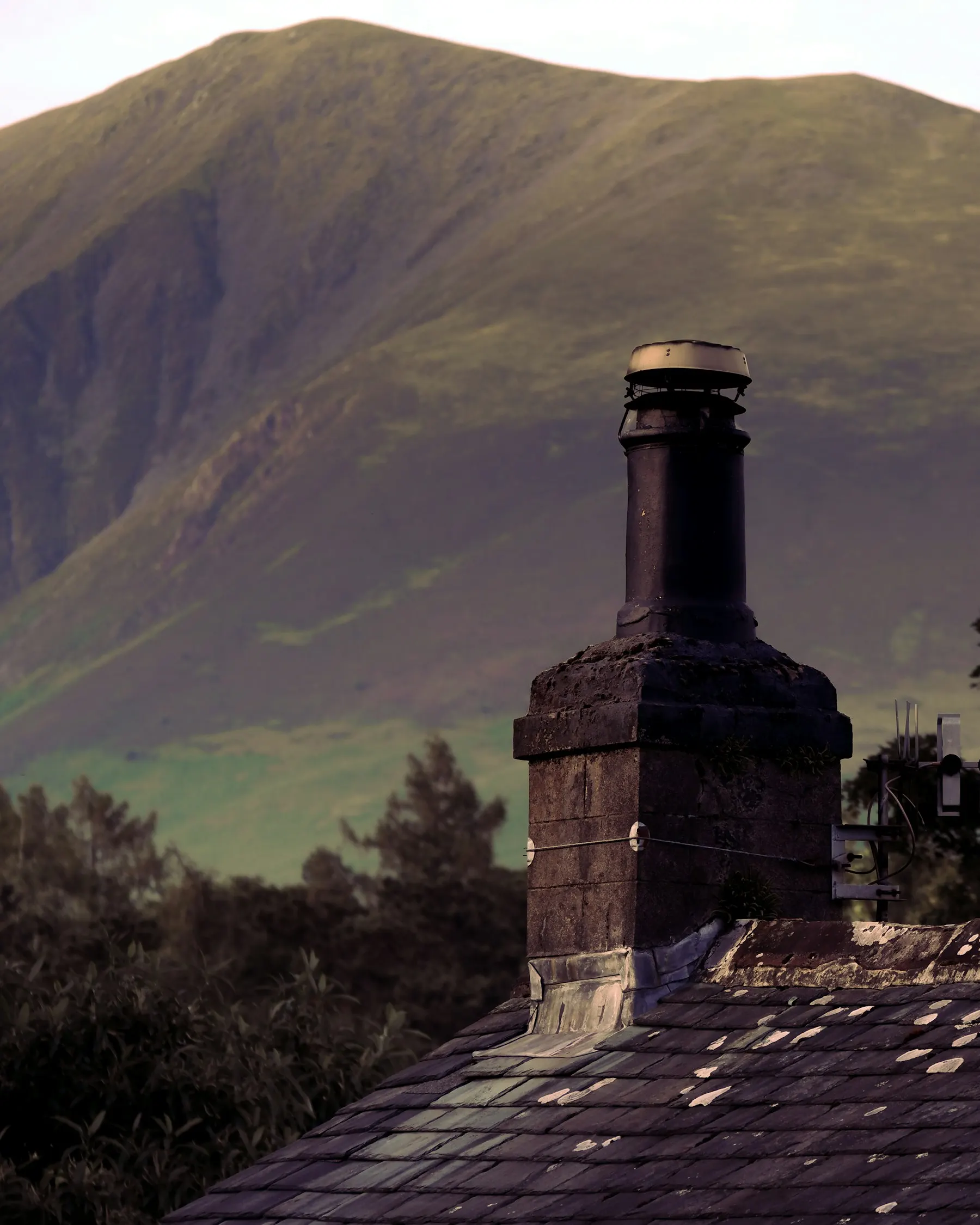 A stone chimney with a cowl on a slate roof, with a fell behind